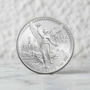 May include: A 1982 Mexican silver coin featuring the Winged Victory statue. The coin is inscribed with 1 ONZA, PLATA PURA, and LEY 999. The design depicts the angel figure with outstretched wings standing before mountains, with the Mexico mint mark.
