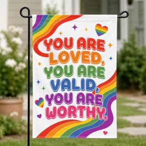 May include: A white garden flag featuring a rainbow-colored wavy border and the text "YOU ARE LOVED, YOU ARE VALID, YOU ARE WORTHY." The design includes small colorful stars and hearts, with a prominent rainbow heart in the upper right corner.