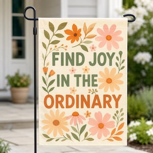 May include: A beige garden flag with the words "FIND JOY IN THE ORDINARY" in green and orange. The flag is decorated with orange, peach, and pink flowers and green leaves. The flag is hanging on a black metal stand.