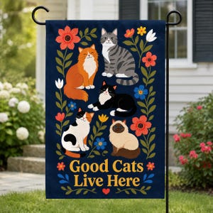 May include: A dark blue garden flag featuring five cats of various colors and patterns surrounded by colorful flowers and green leaves. The text Good Cats Live Here is printed in yellow at the bottom, accompanied by a small red heart icon.