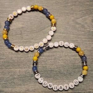 May include: Two beaded bracelets featuring white letter beads spelling IRINA FOUNDATION. The bracelets are composed of alternating blue, yellow, and clear beads, accented with gold-toned and silver-toned metallic spacers throughout the circular designs.