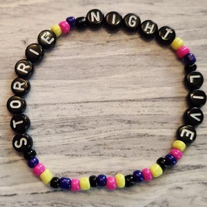 May include: A circular beaded bracelet featuring black letter beads that spell out STORRIE NIGHT LIVE. The design includes a repeating pattern of small, round, colorful beads in shades of yellow, pink, blue, and black interspersed throughout.