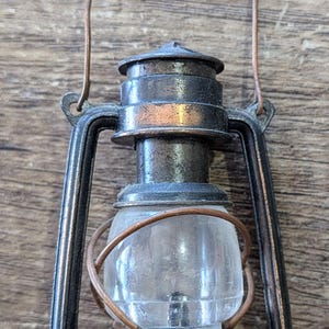May include: A miniature lantern with a dark, weathered bronze-toned metal frame and a clear, rounded glass globe. The lantern features a thin wire handle at the top, a circular base with a side opening, and a protective wire cage around the glass.