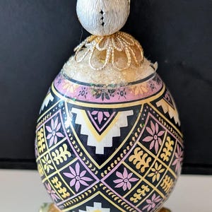 May include: A decorative egg ornament featuring a black, pink, white, and gold geometric and floral pattern. The egg is topped with a white finial and sits on a gold-toned metal stand with three curved legs, showcasing intricate detail.