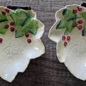 Könnte beinhalten: Zwei Keramikschalen in Form von cremefarbenen Blättern mit geprägten Blattadern. Jede Schale hat einen dunkelbraunen Stiel am oberen Rand, grün bemalte Blätter und kleine rote Beeren. Die Oberfläche der Schalen weist eine feine Craquelé-Glasur auf.