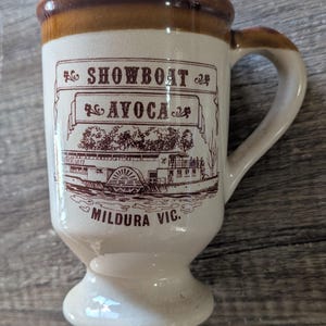 May include: A cream-coloured ceramic mug with a brown rim and handle. The front features a brown graphic of a paddle steamer boat with the text SHOWBOAT AVOCA and MILDURA VIC. The mug has a pedestal base and a smooth, glossy finish throughout.