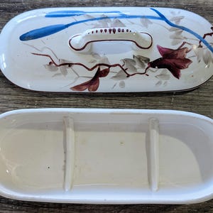May include: An oval ceramic butter dish with a lid featuring a central handle. The lid is decorated with hand-painted blue, brown, and dark red floral motifs on a white background. The base is a plain white oval tray with two interior dividers.