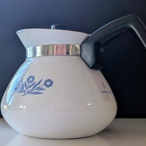 May include: A white ceramic teapot featuring a blue floral pattern on the side, a black handle, and a black lid. A silver-coloured metal band encircles the upper portion of the pot. The text CORNING WARE is printed in small black letters on the side.