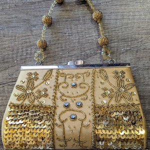 May include: A gold-toned handbag featuring a beaded handle with spherical accents. The exterior is decorated with gold sequins, intricate beadwork in floral patterns, and a central panel with small blue stones and linear bead embellishments.