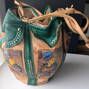 Könnte beinhalten: Eine Beuteltasche aus hellbraunen und dunkelgrünen Lederbahnen. Die Tasche verfügt über Ziernähte und rechteckige Einsätze, die eine ägyptische Figur in Blau und Gelb darstellen. Sie umfasst einen hellbraunen Lederriemen und einen Kordelzug.
