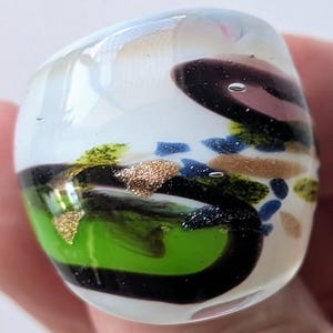 May include: A chunky glass ring featuring a clear base with swirling patterns of black, bright green, and tan. The design includes embedded gold-coloured glitter flecks and small air bubbles trapped within the smooth, rounded, polished glass surface.