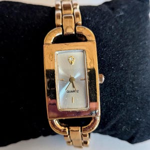 Puede incluir: Un reloj de cuarzo rectangular en tono dorado con esfera plateada de efecto rayos de sol, una piedra transparente en la posición de las doce y una pulsera de eslabones de metal pulido. La esfera muestra la palabra QUARTZ en letras negras.