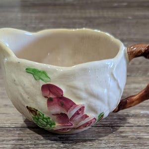 May include: Cream ceramic creamer with a brown handle and floral design. The creamer has a spout and is decorated with red and green floral accents. The handle has a textured, brown appearance.