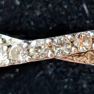May include: A silver-toned crossover ring featuring two intersecting bands. Each band is encrusted with a continuous row of small, round, clear, faceted stones that catch the light, creating a sparkling effect across the entire surface of the ring.