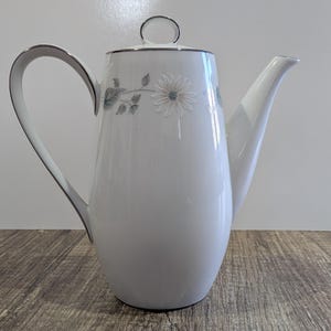 May include: White ceramic coffee pot with a silver rim and handle. The pot features a floral design with a daisy and leaves. The spout is long and curved. The lid has a silver ring handle.