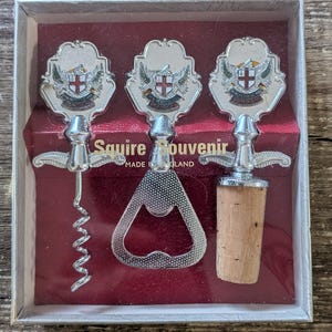 May include: A Squire Souvenir set made in England featuring three silver-toned metal tools: a corkscrew, a bottle opener, and a wine stopper with a cork base. Each tool has a decorative crest emblem with a red cross and the text London.