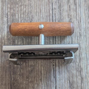 May include: A kitchen tool featuring a horizontal wooden handle attached to a silver-coloured metal base. The base has a serrated edge and a U-shaped frame with notched sides, designed for gripping or cutting, with a single metal rivet in the handle.