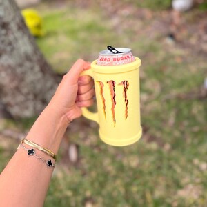 May include: A yellow mug-shaped can cooler with a handle, holding a Monster Energy drink can. The cooler has three claw-like cutouts and the words "ZERO SUGAR" are visible on the can. The background is blurred with green grass and a tree.