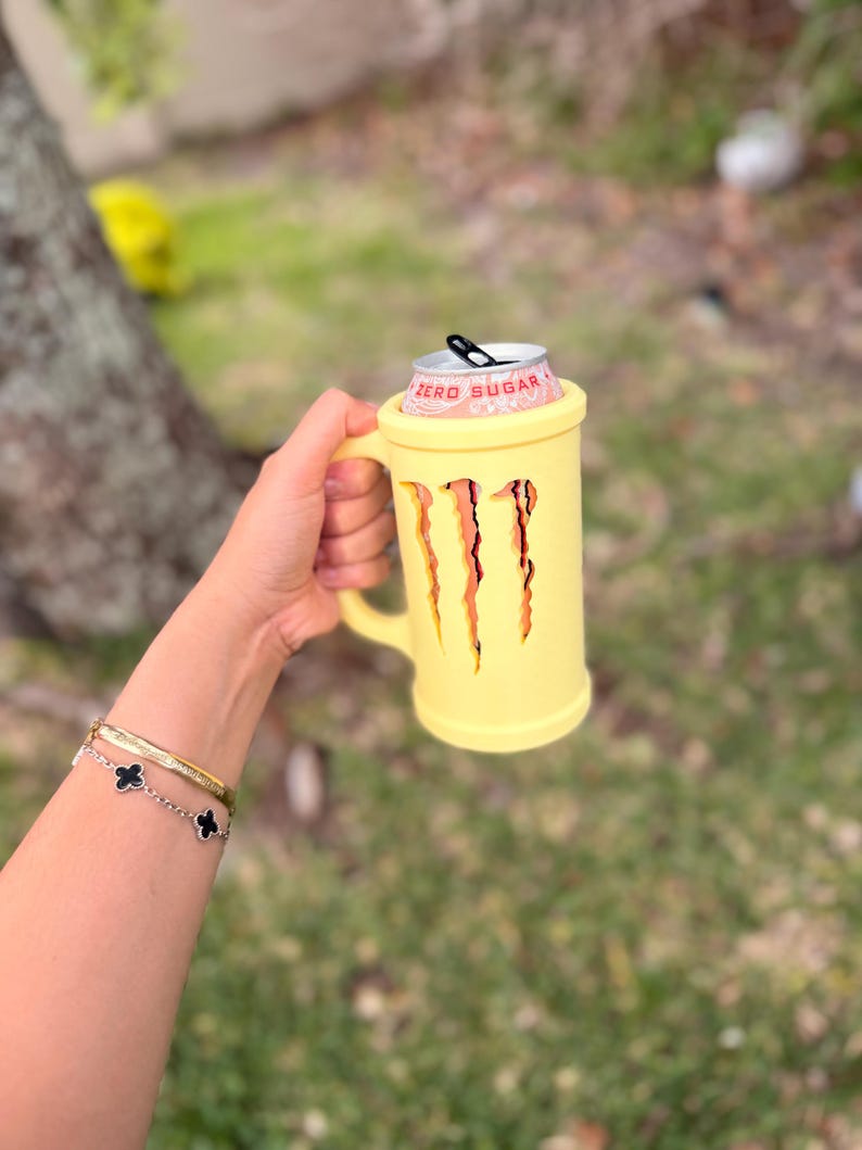 May include: A yellow mug-shaped can cooler with a handle, holding a Monster Energy drink can. The cooler has three claw-like cutouts and the words "ZERO SUGAR" are visible on the can. The background is blurred with green grass and a tree.