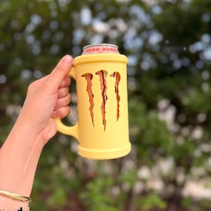May include: A yellow mug with the Monster Energy logo cut out. The mug holds a Monster Energy Zero Sugar can. The mug has a handle and is held by a person. The background is blurred green foliage.