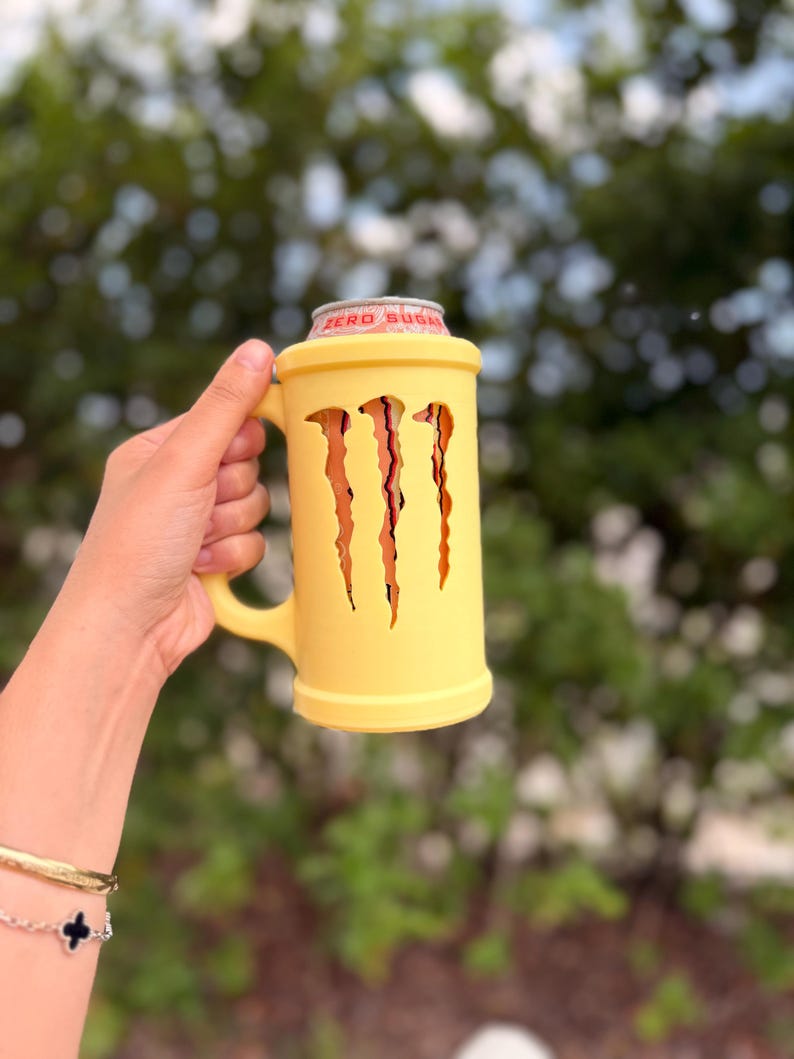 May include: A yellow mug with the Monster Energy logo cut out. The mug holds a Monster Energy Zero Sugar can. The mug has a handle and is held by a person. The background is blurred green foliage.
