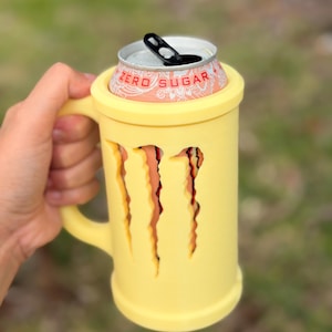 May include: A yellow mug-shaped can cooler with the Monster claw marks cut out. A Monster Energy Zero Sugar can sits inside the cooler. The cooler has a handle and is held by a person.