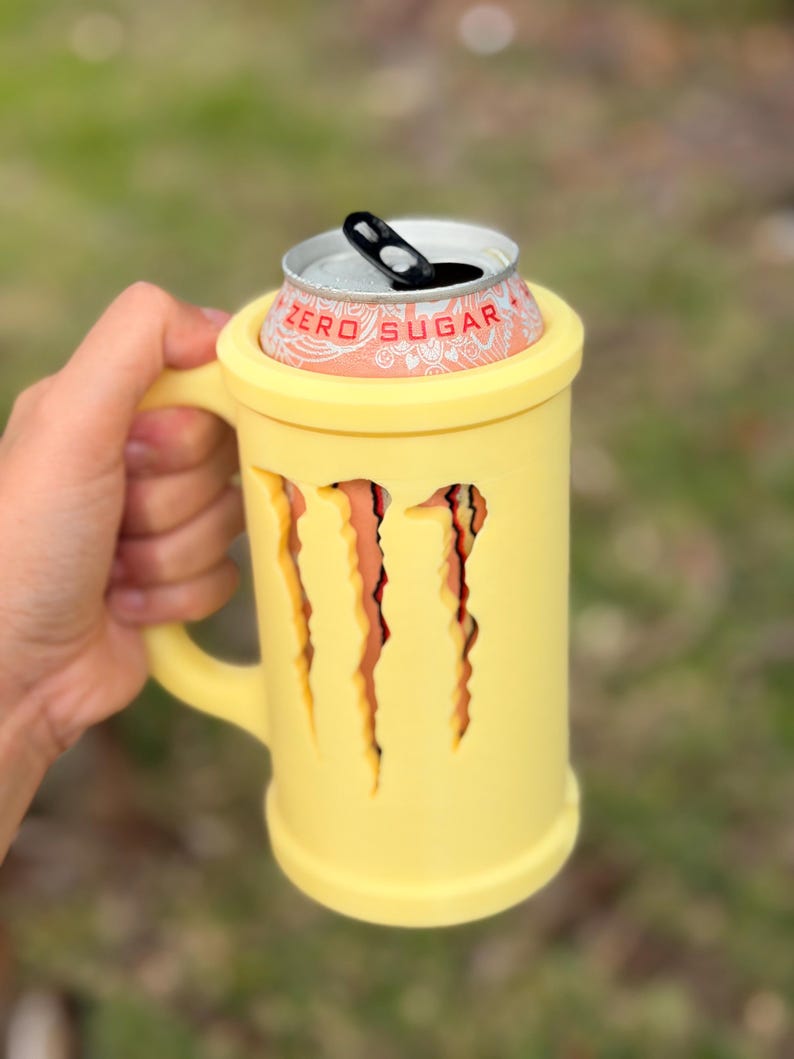 May include: A yellow mug-shaped can cooler with the Monster claw marks cut out. A Monster Energy Zero Sugar can sits inside the cooler. The cooler has a handle and is held by a person.