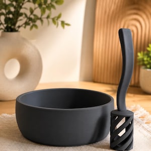 May include: A matte charcoal gray circular bowl with smooth, high walls and a matching charcoal gray kitchen tool featuring a long, curved handle and a cylindrical, spiraled, open-work head designed for mixing or whisking tasks.