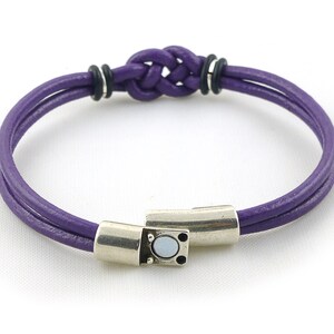 Puede incluir: Un brazalete de cuero morado con un cierre de plata y un detalle de nudo. El brazalete est&aacute; hecho de dos hebras de cuero trenzadas juntas.