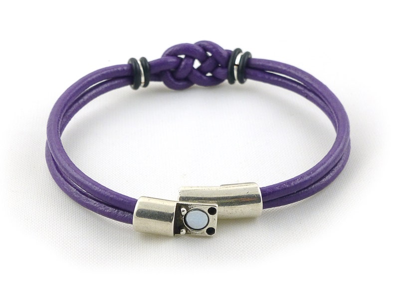 Puede incluir: Un brazalete de cuero morado con un cierre de plata y un detalle de nudo. El brazalete est&aacute; hecho de dos hebras de cuero trenzadas juntas.