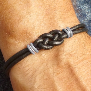 May include: A black leather bracelet with a nautical knot design. The bracelet features two parallel leather cords and is accented with silver-colored metal rings. The bracelet is shown on a wrist.