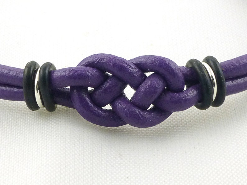 Puede incluir: Un brazalete de cuero morado con un dise&ntilde;o de nudo y detalles en negro y plata.