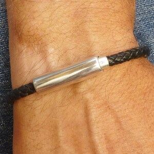 May include: Black braided leather bracelet with a silver-colored cylindrical clasp. The bracelet is worn on a wrist, with a denim background visible.