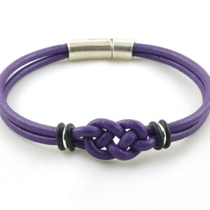 Puede incluir: Un brazalete de cuero morado con un dise&ntilde;o de nudo y cierre magn&eacute;tico plateado. El brazalete tiene dos anillos negros a cada lado del nudo.