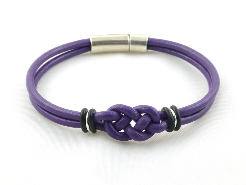 Puede incluir: Un brazalete de cuero morado con un dise&ntilde;o de nudo y cierre magn&eacute;tico plateado. El brazalete tiene dos anillos negros a cada lado del nudo.
