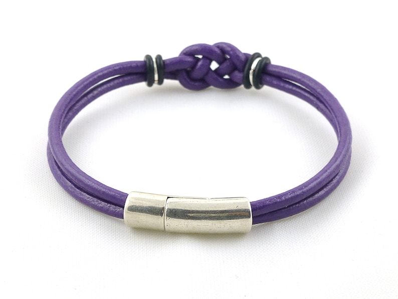 Puede incluir: Un brazalete de cuero morado con un cierre magn&eacute;tico plateado y un dise&ntilde;o de nudo trenzado.