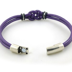 Puede incluir: Un brazalete de cuero morado con un cierre magn&eacute;tico plateado y un dise&ntilde;o de nudo trenzado.