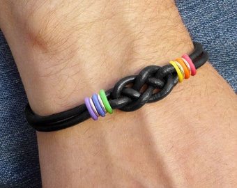 May include: Black leather bracelet featuring a knotted design with colorful accent rings. The bracelet has a thick, braided appearance and is adorned with rings in various colors, including purple, green, silver, yellow, orange, and pink. The bracelet is shown on a wrist.