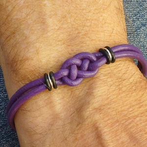 Puede incluir: Un brazalete de cuero morado con un dise&ntilde;o de nudo y dos anillos negros y plateados.