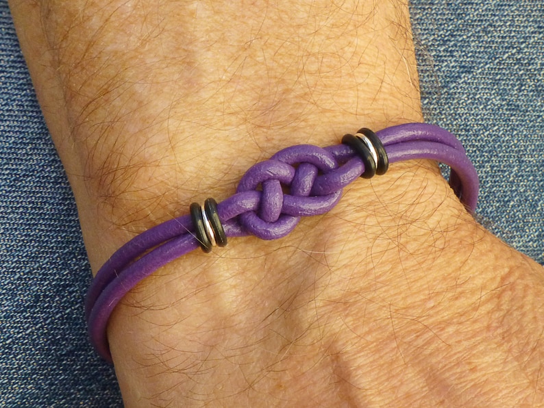 Puede incluir: Un brazalete de cuero morado con un dise&ntilde;o de nudo y dos anillos negros y plateados.