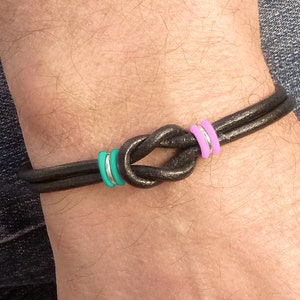 May include: A black leather bracelet with a knotted design and two accent rings, one pink and one turquoise.