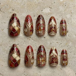 May include: A set of ten press-on nails featuring a stained glass design in shades of deep red, cream, and gold. Each nail has a pointed almond shape with intricate gold lines forming geometric patterns and arched window-like motifs.