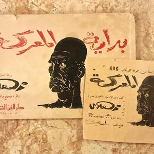 May include: Two rectangular paper prints featuring a black ink illustration of a stern-faced person in profile. The prints contain Arabic text in red and black ink, including the phrase "بداية المعركة" and "دار الفن الحديث" printed on a light beige background.