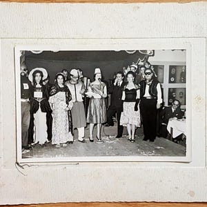 May include: A black and white vintage photograph mounted on a textured white card frame, depicting a group of people in various costumes standing on a stage. The individuals are dressed in formal attire, masks, and hats, with one person holding a microphone.