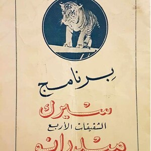 May include: A vintage paper poster featuring a dark blue circular emblem with a tiger standing on a ledge. Below the emblem, the text is written in Arabic script in dark blue and red ink, enclosed within a thin rectangular border on cream paper.