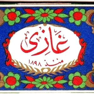 Puede incluir: Una etiqueta rectangular de caja de cerillas con un óvalo blanco central que contiene escritura árabe roja y el año 1898. El borde presenta un fondo azul vibrante decorado con un patrón repetitivo de flores verdes y pétalos rojos con detalles dorados.