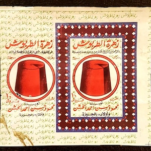 May include: A square paper label featuring two identical red fez hat illustrations inside circular frames. The design includes repeating Arabic text in the background, a dark red and blue geometric border, and bold red Arabic script text.