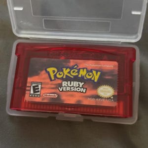May include: A red Game Boy Advance cartridge for the game "Pokémon Ruby Version" is inside a clear plastic case. The cartridge has a label with the Pokémon logo and game title. The case is open.