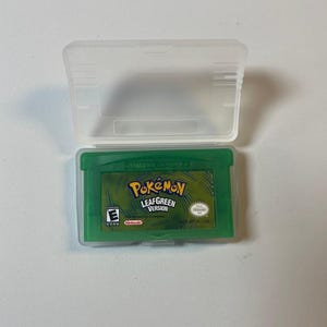 Peut inclure: Une cartouche verte de Game Boy Advance pour le jeu "Pokémon LeafGreen Version" est présentée dans un étui en plastique transparent. La cartouche de jeu a une étiquette verte avec le logo du jeu et la classification ESRB.