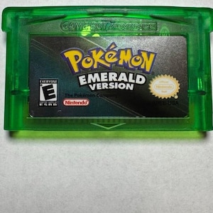 May include: A green Game Boy Advance cartridge for the game "Pokémon Emerald Version." The cartridge has a black label with the game title in yellow and white, and the Nintendo logo. The ESRB rating is visible.